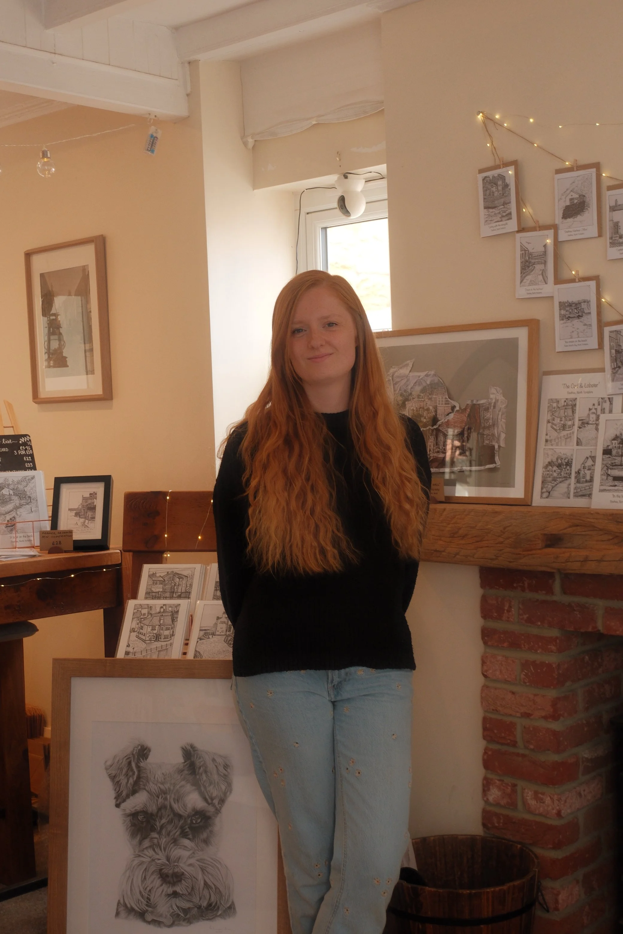 A young woman with long red hair standing indoors near artwork and framed pictures on display. She is wearing a black top and light blue jeans. The background includes a window, fairy lights, and art prints on the wall.