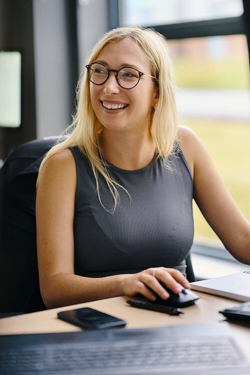 reportage style team headshot of woman with long blonde hair and glasses sat smiling at desk