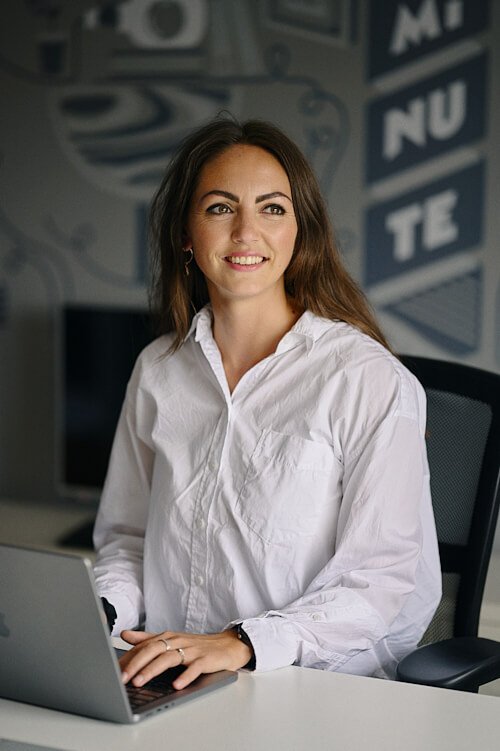 reportage style team headshot of smiling young woman with long dark hair and white shirt sat at a laptop