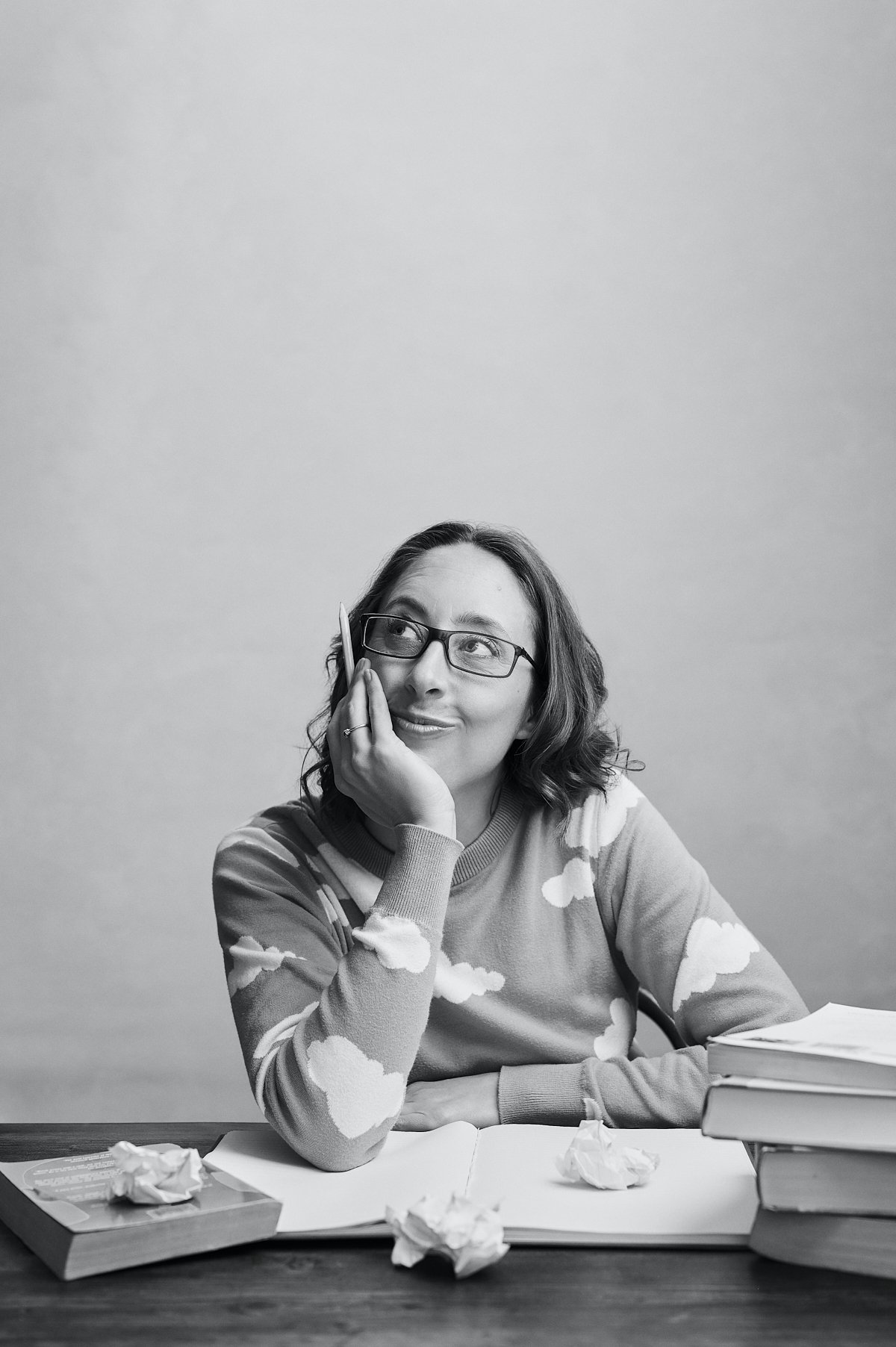 Black and white Portrait of Maz Evans sitting at her desk surrounded by scrunched up paper and a pile of books