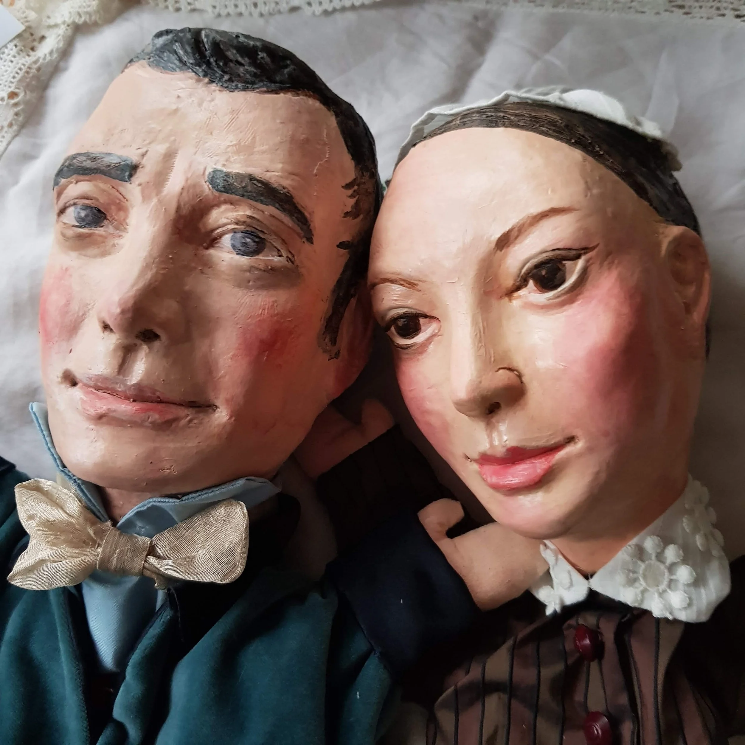 Two vintage-style puppets of a man and a woman with detailed facial features, positioned close together. The man puppet has dark hair, thick eyebrows, and is wearing a suit with a bow tie. The woman puppet has dark hair, a white headscarf, and a dark