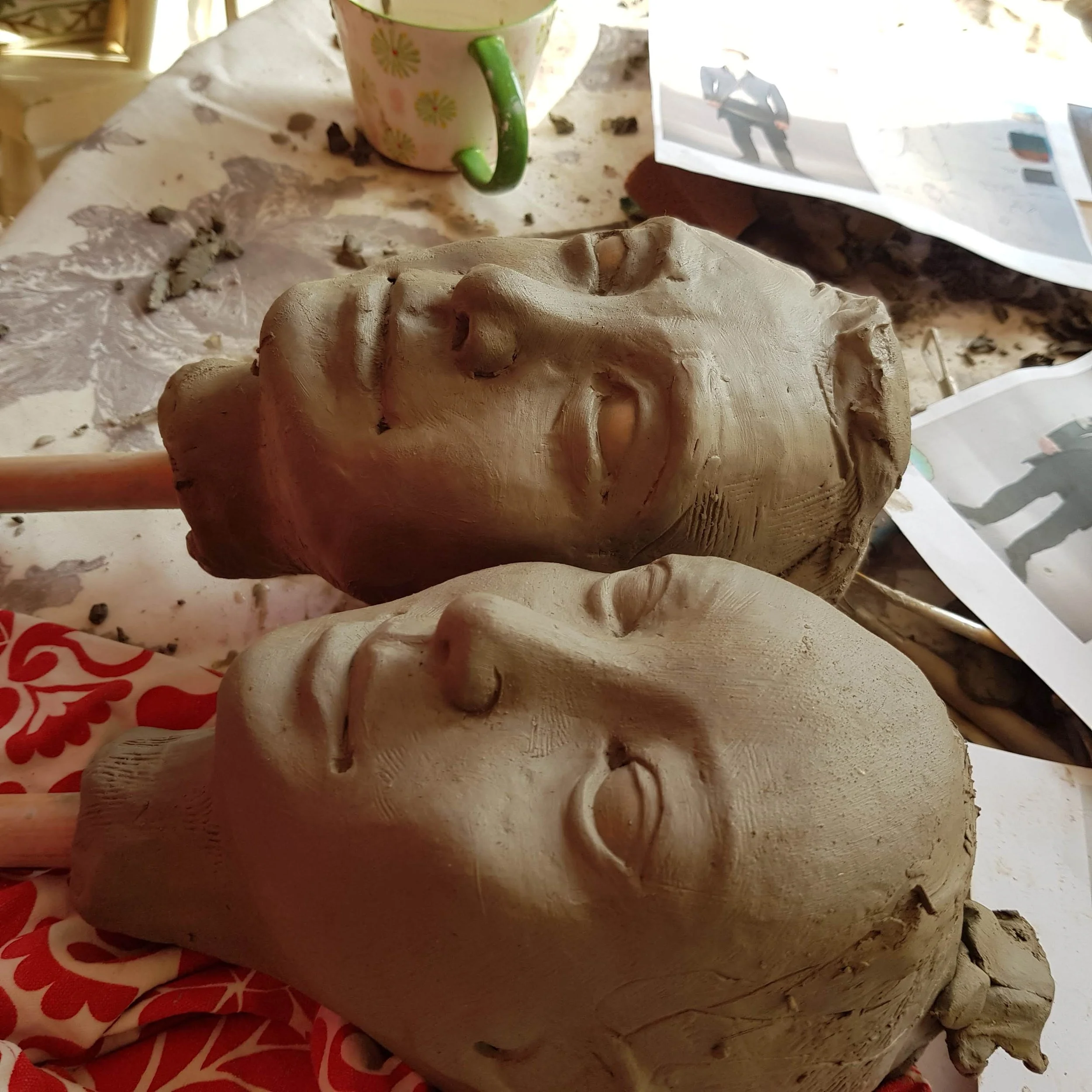 Two unfinished clay sculptures of human faces lying on their sides on a table, surrounded by sculpting materials and papers.