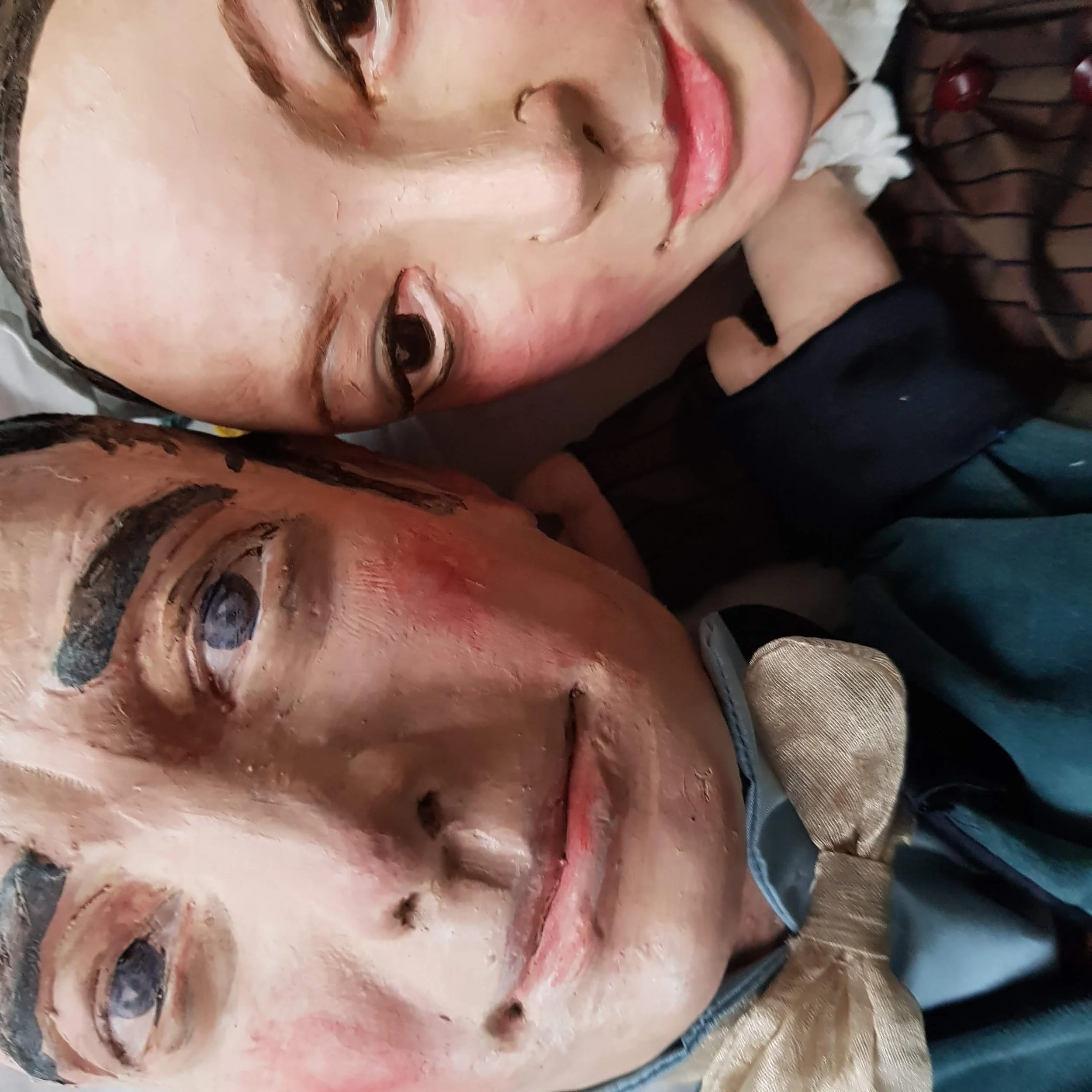 Close-up of two realistic, painted masks resembling human faces, one with short hair and a nose piercing, the other with slightly closed eyes and a faint smile, layered closely together.