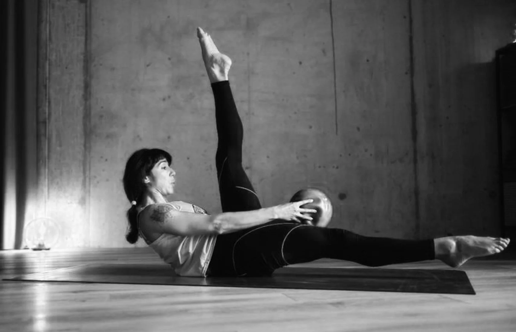 A woman practicing yoga indoors, lying on a mat with one leg extended and the other leg raised while holding a ball, in black and white.
