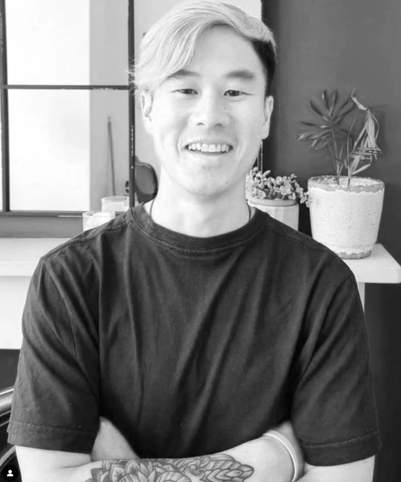 Black and white photo of a young man with light-colored hair smiling, wearing a dark T-shirt with his arms crossed, standing in front of a window and a table with potted plants.