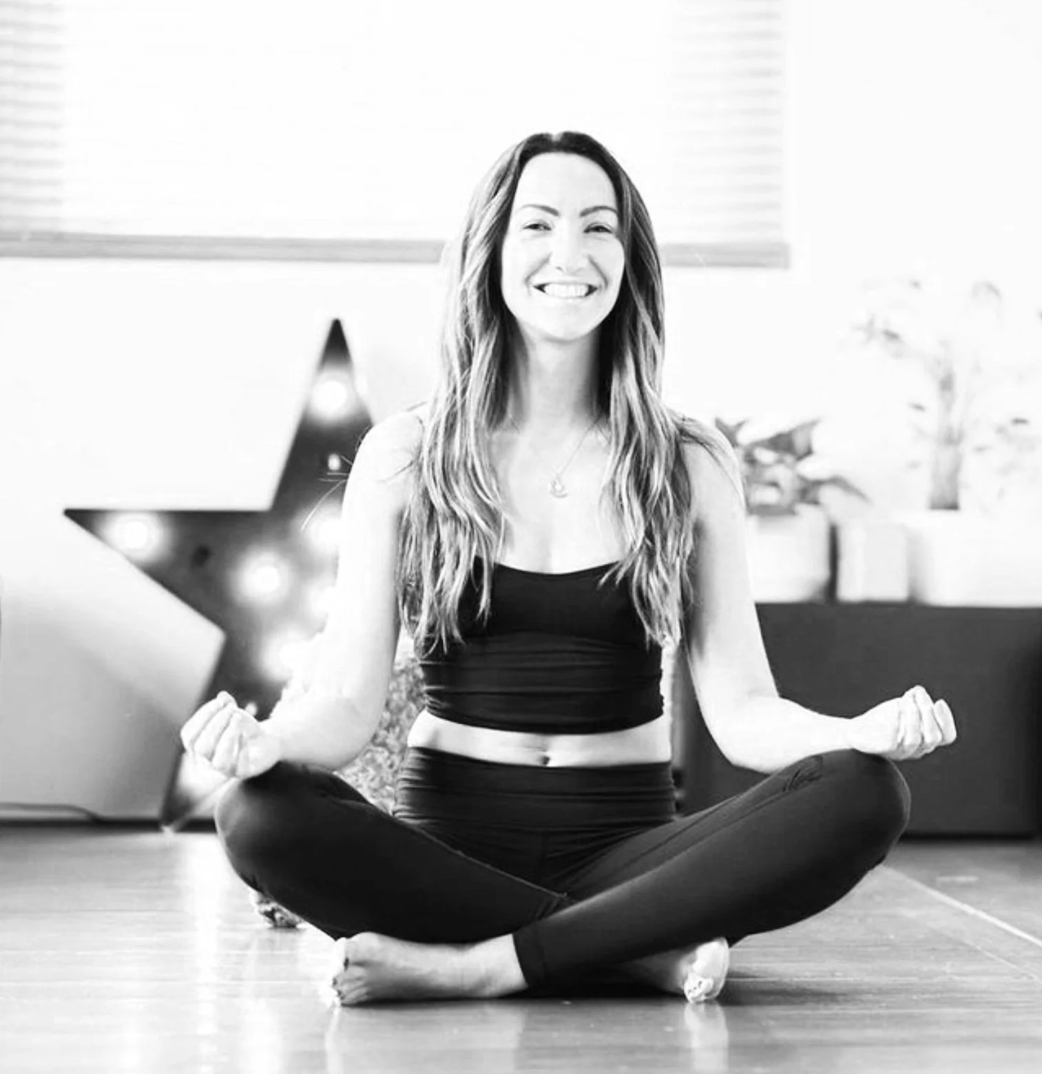 A woman sitting in a cross-legged yoga pose indoors, smiling, with a decorative star and plants in the background.