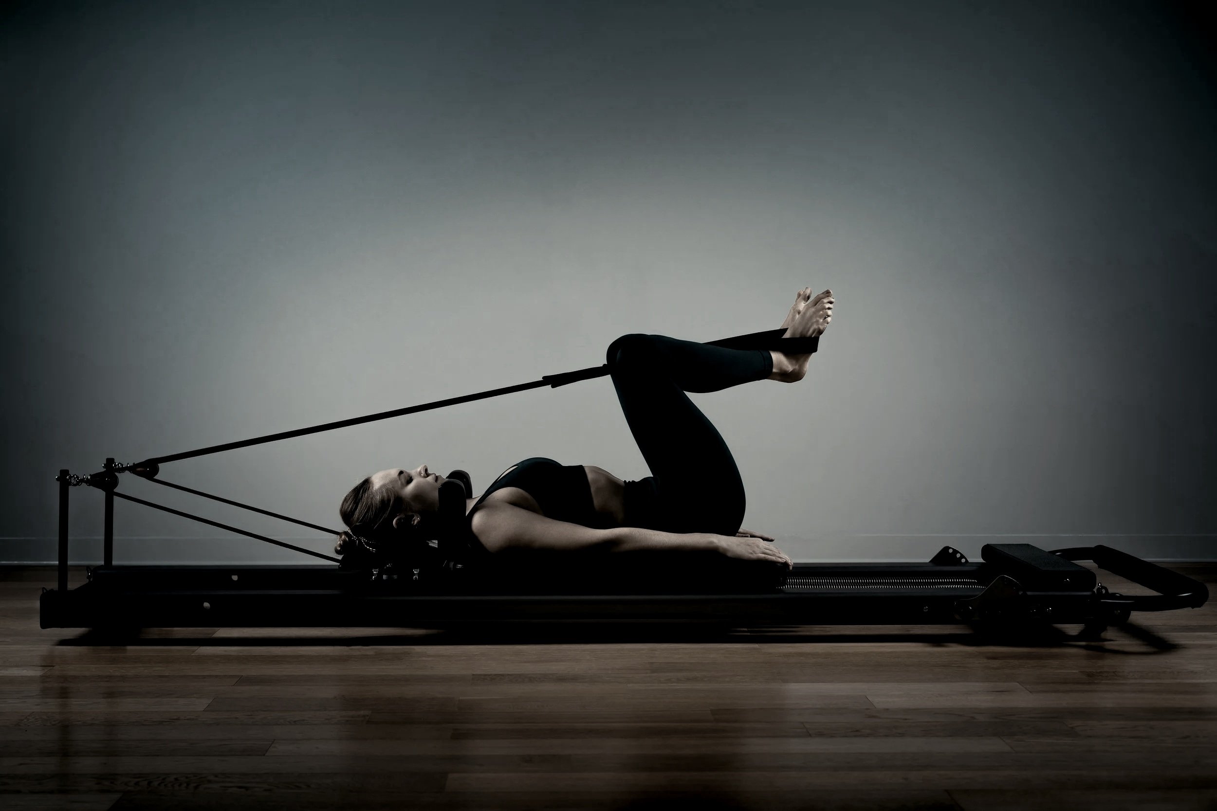 A woman practicing yoga indoors, lying on a mat, holding a blue exercise ball with her legs raised straight in the air, against a concrete wall background.