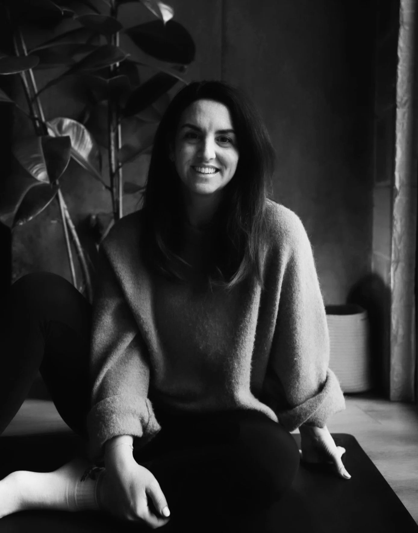 A smiling woman with long dark hair, sitting cross-legged on a yoga mat indoors, with houseplants and textured walls in the background. Black and white photo.