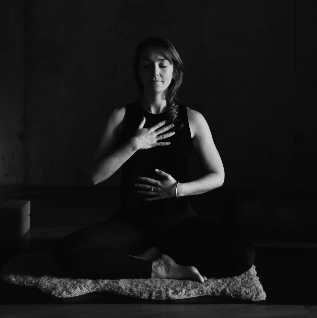 A woman sitting cross-legged on a rug, practicing meditation with her eyes closed and hands on her chest and abdomen, in a dark room.