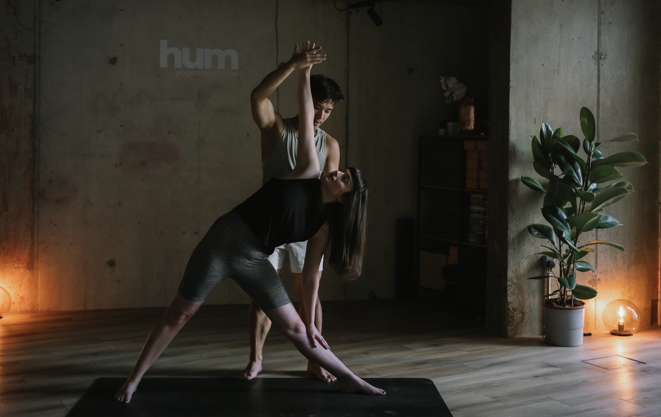 A man and woman practicing yoga indoors, with the woman in a side bend pose and the man supporting her, in a room with a potted plant and warm lighting.