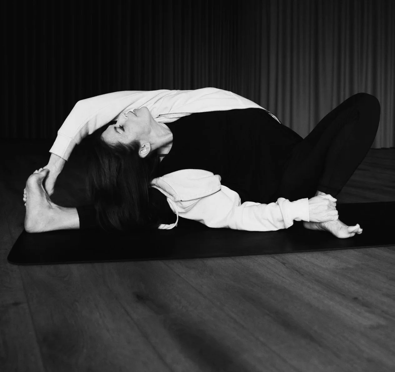 A woman practicing yoga indoors, lying on a yoga mat, performing a stretch with one leg bent, the other extended, and arms reaching over her head, dressed in black and white athletic clothing.