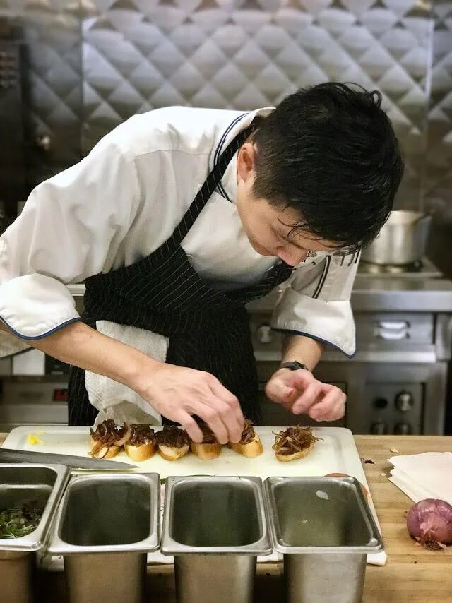 Chef preparing small canapés with caramelized onions on a cutting board in a professional kitchen.