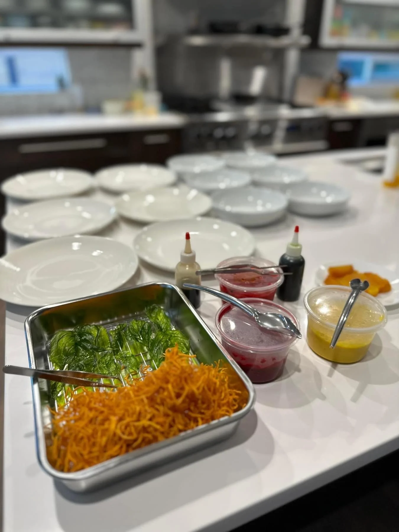 A kitchen counter with several white plates, a metal tray with fresh green leaves and shredded orange carrots, small bottles of sauces, and bowls of red and yellow sauces. The background shows a stove and kitchen shelves.