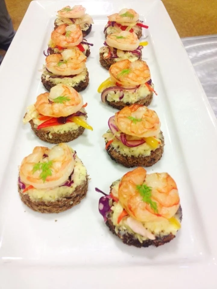 Appetizer bites with round, dark bread bases topped with cheese, shrimp, and colorful sliced vegetables on a white rectangular platter.