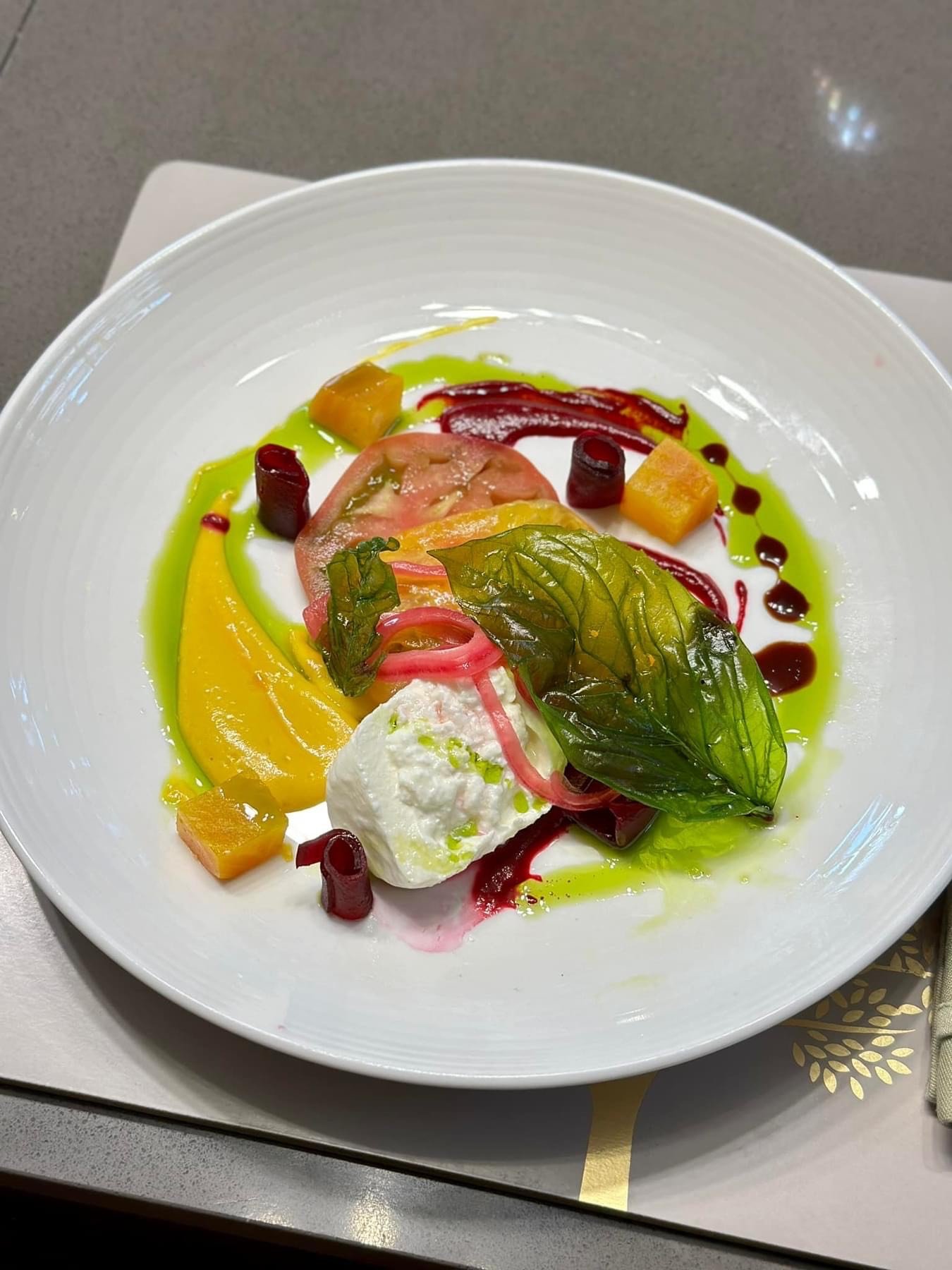 A colorful salad on a white plate containing mixed greens, yellow sauce, beetroot, tomato, melon, and a dollop of white cheese, garnished with drizzles of red and green sauces.