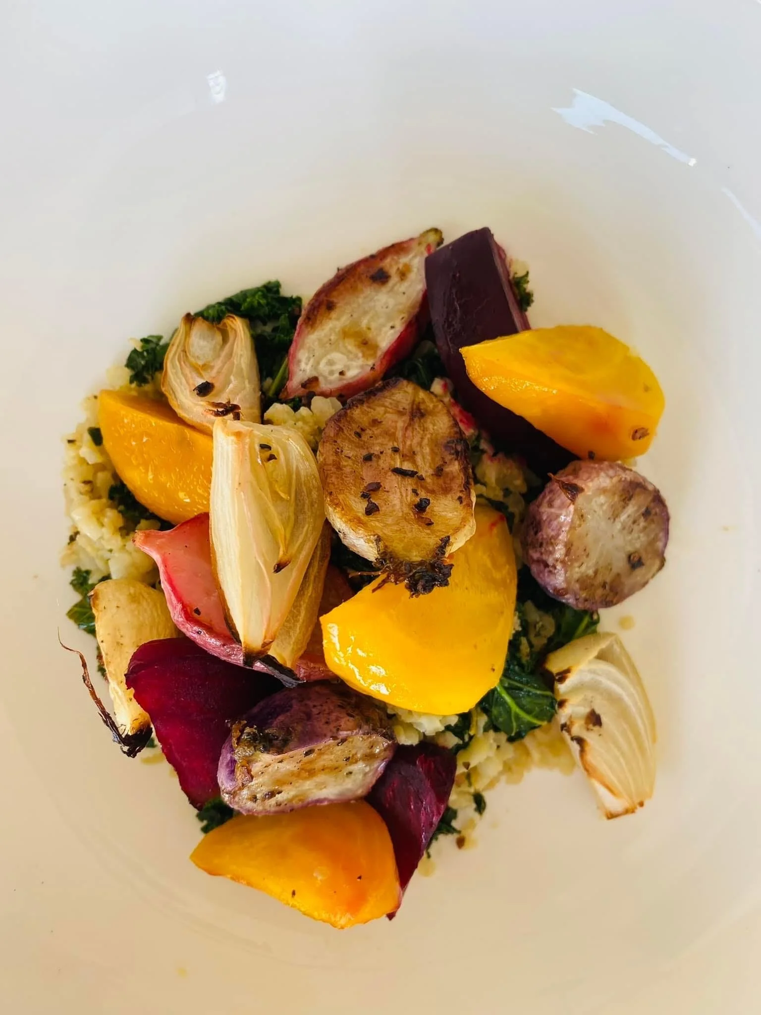 Roasted vegetables including yellow, red, purple potatoes, artichoke hearts, and greens in a white bowl.