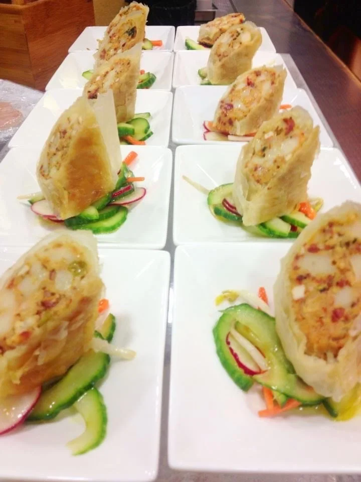 Multiple servings of egg rolls topped with sliced cucumbers, radishes, and shredded carrots on white plates.