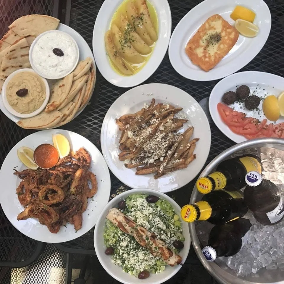 A table filled with traditional Greek dishes including grilled pita bread, tzatziki, hummus, sliced melon, fried fish, grilled chicken salad, grilled meatballs, and fried squid, alongside bottles of beer on ice.