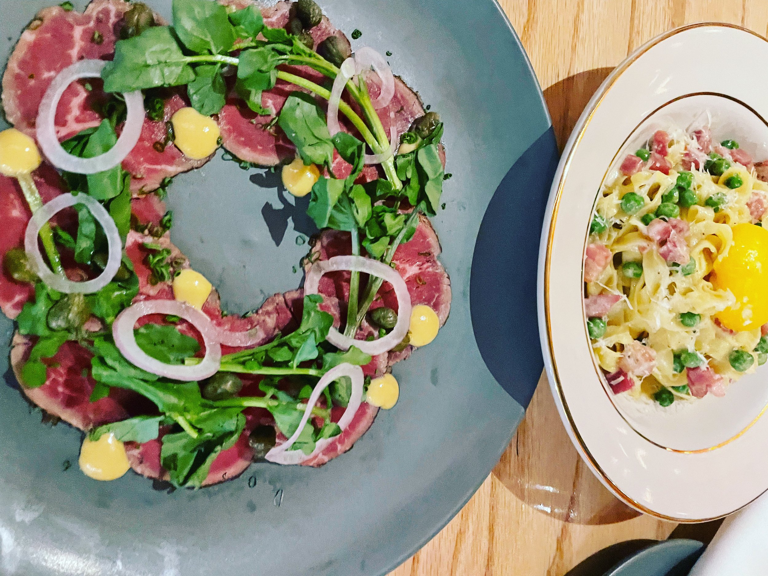 Crempeta with thinly sliced raw beef, arugula, capers, onion rings, and yellow sauce, and a bowl of pasta with peas, ham, and a raw egg yolk on top.