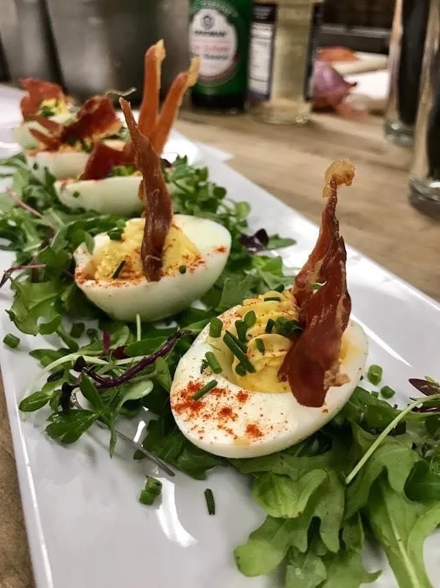 Deviled eggs garnished with chopped chives, paprika, and topped with crispy bacon strips, arranged on a bed of mixed greens on a white serving platter.