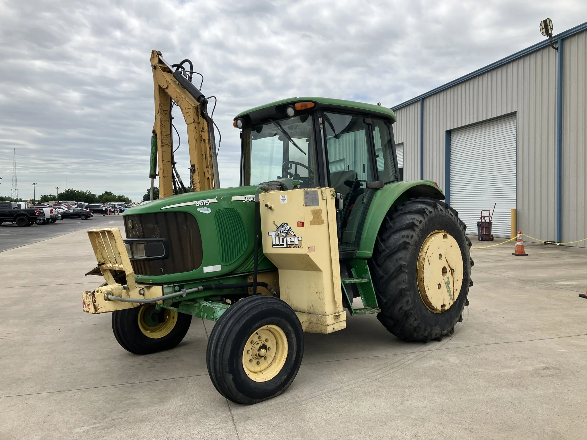 Green tractor with yellow attachments parked outside near a large industrial building with a blue roof and rolled-up doors.