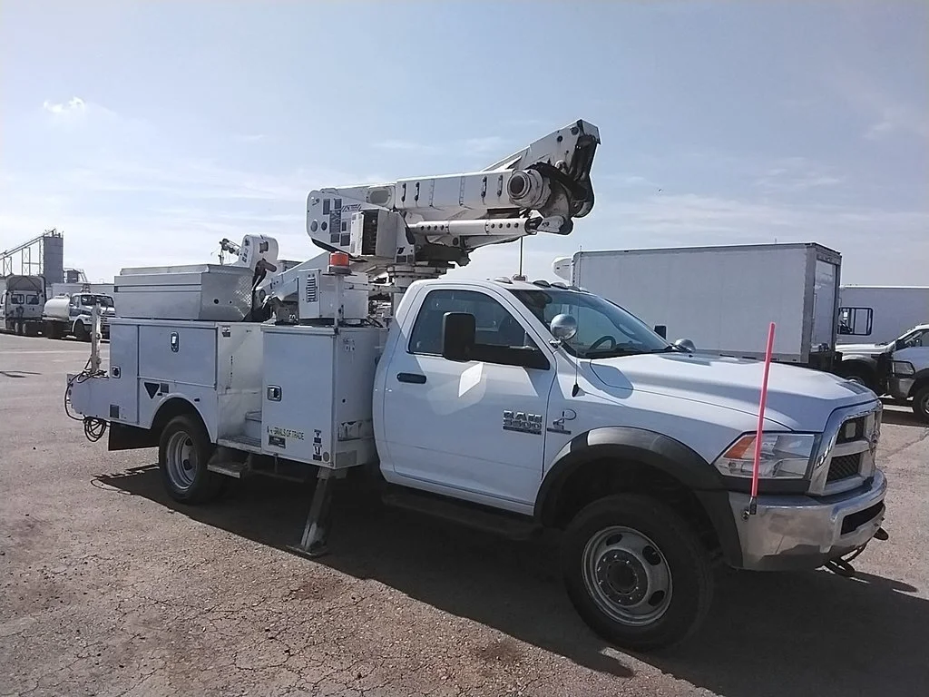 A white utility truck equipped with a bucket lift, parked in a lot with other trucks and vehicles.