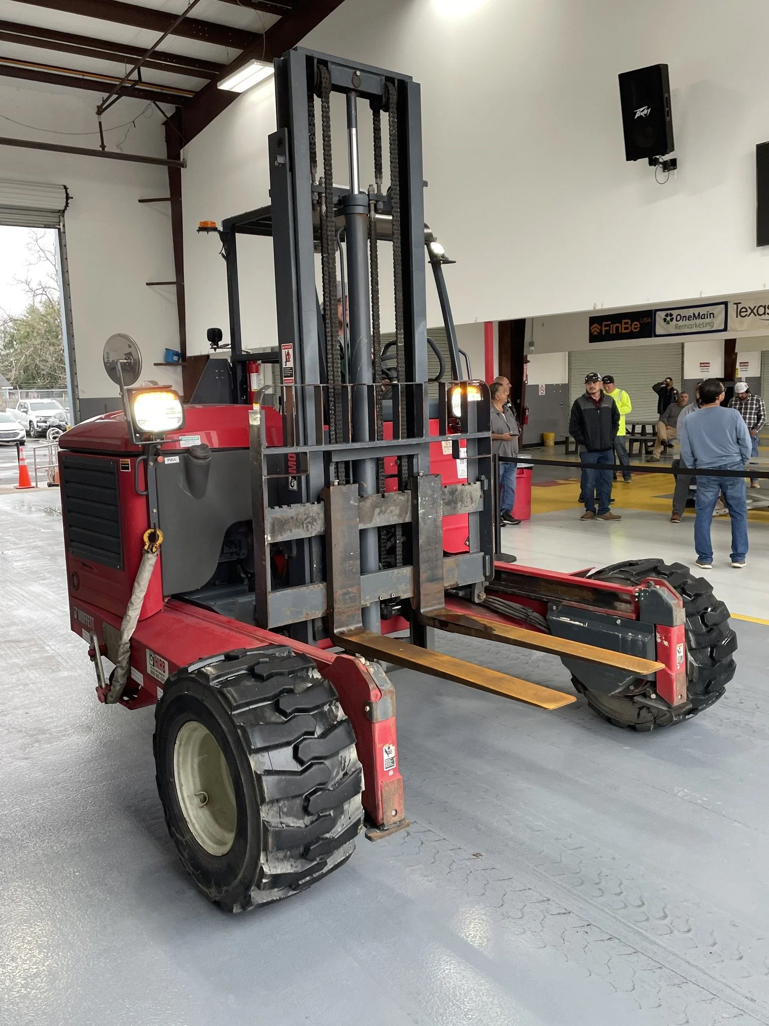 A red forklift is inside a large garage or warehouse with group of people in the background, some wearing safety vests, and signs on the wall.