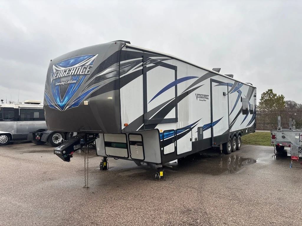 A Vengeance gaming edition travel trailer parked on a wet lot, with a white vehicle and other equipment nearby, under a cloudy sky.