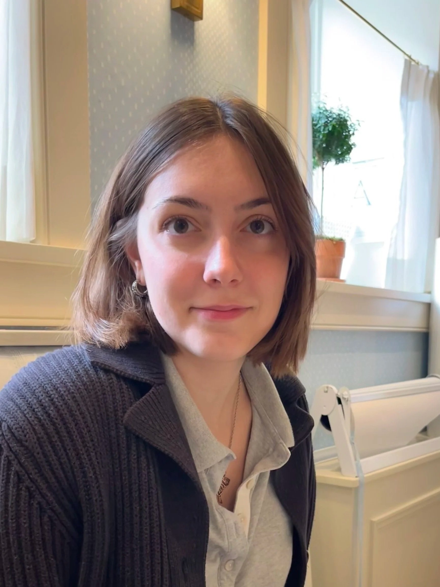 A young woman with shoulder-length brown hair and earrings, sitting indoors in a room with wallpapered walls, a window with white curtains, and a potted plant in the background.
