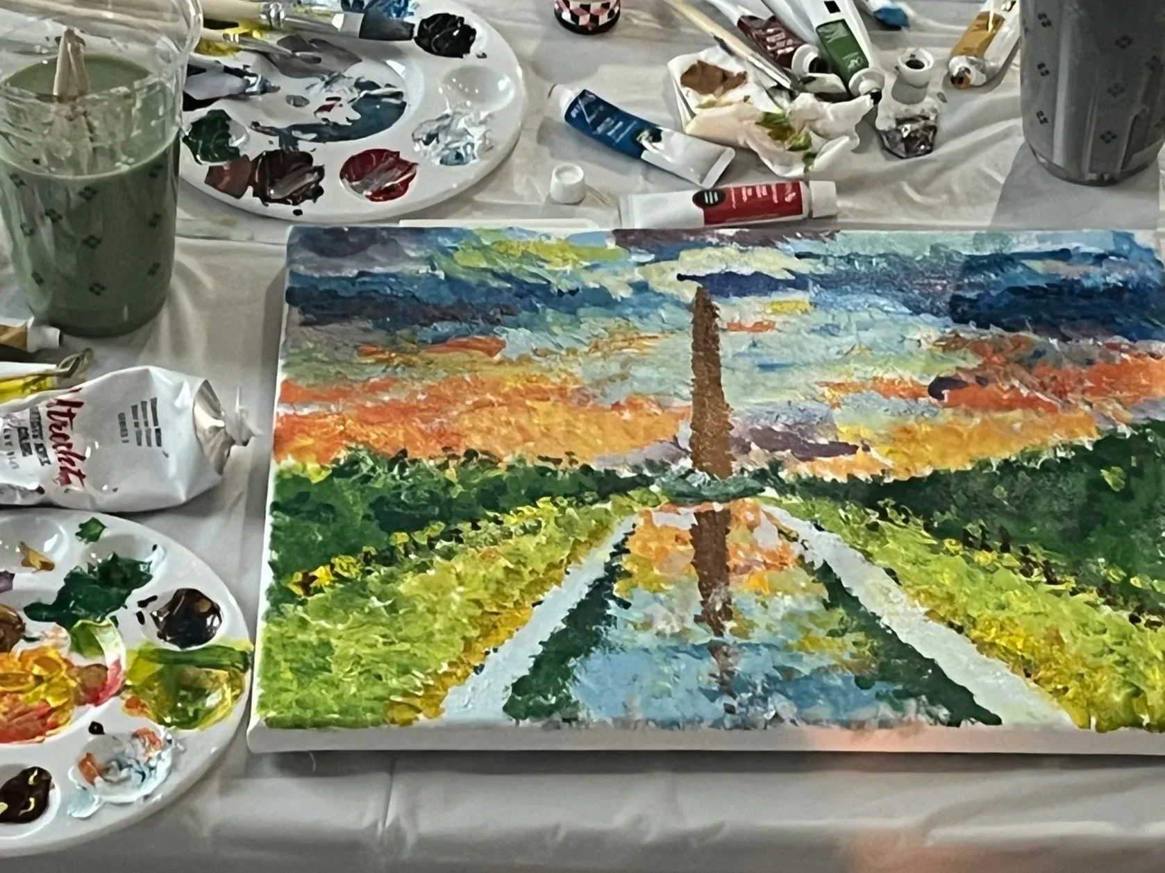 Impressionist painting class. An art workspace with a painting of the Washington Monument at sunset, surrounded by painting supplies including brushes, tubes of paint, palettes, and cups of water.