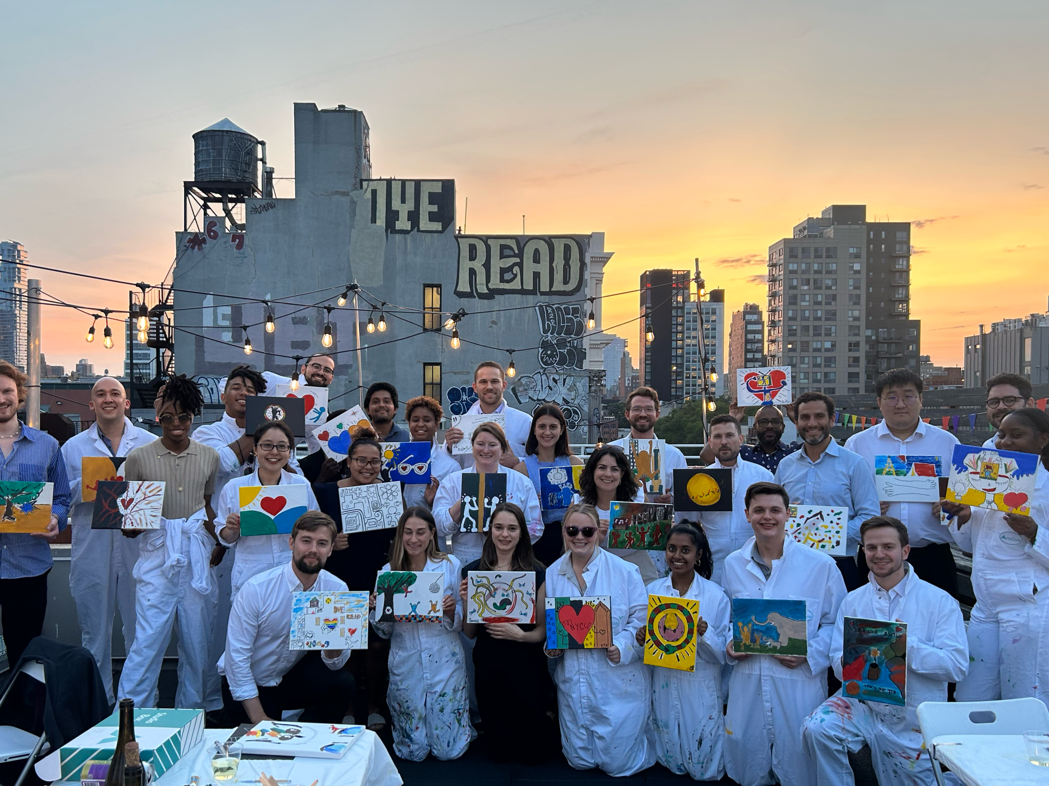 Group of people on a rooftop at sunset, holding up paintings they created, surrounded by city buildings and string lights.