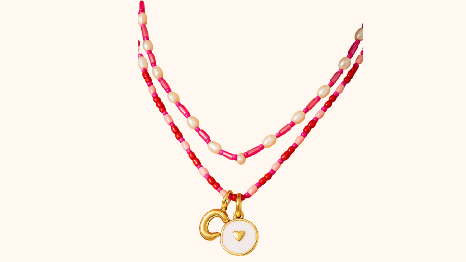 Two layered beaded necklaces with gold charms, one with a heart inside a circle and the other with a letter 'C', against a light background.