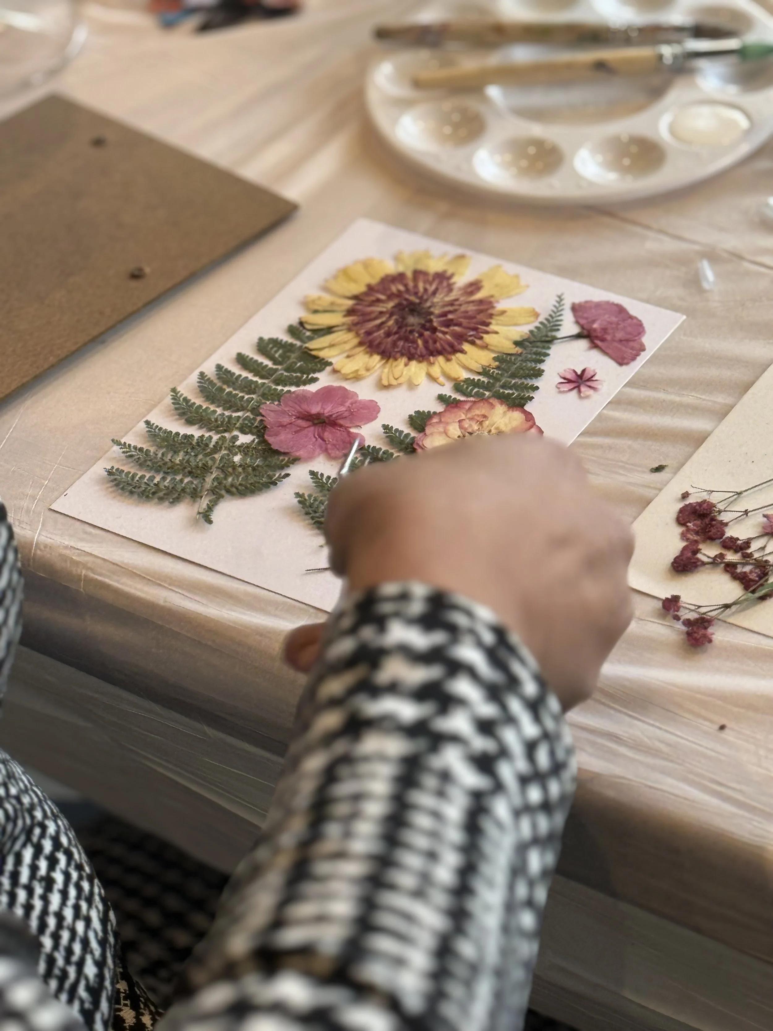 Person arranging pressed flower artwork on table with art supplies nearby.