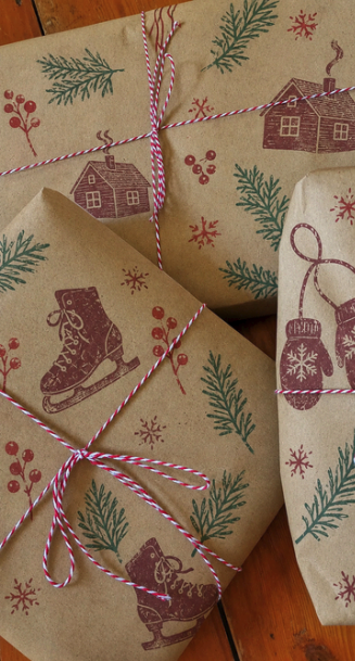 Christmas wrapped gifts decorated with winter-themed stamps of houses, pine branches, gloves, ice skates, and snowflakes, tied with red and white striped string.