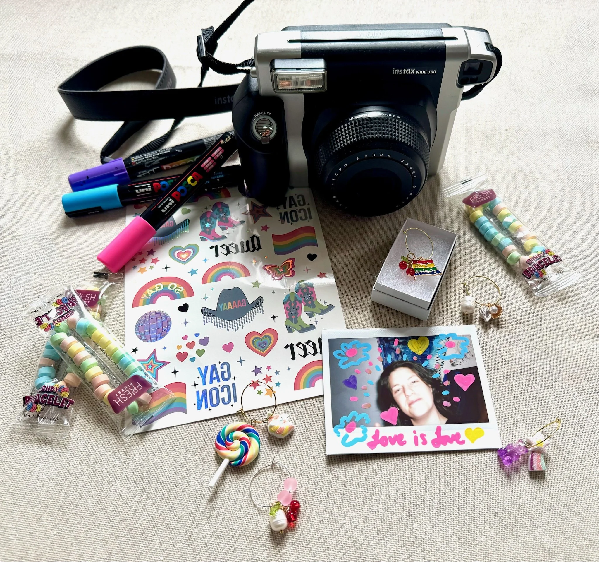 A vintage Instax camera, colorful gel pens, rainbow stickers, a photo of a person with doodles and the caption "Love is love," rainbow earrings, and rainbow-themed accessories scattered on a light-colored fabric surface.