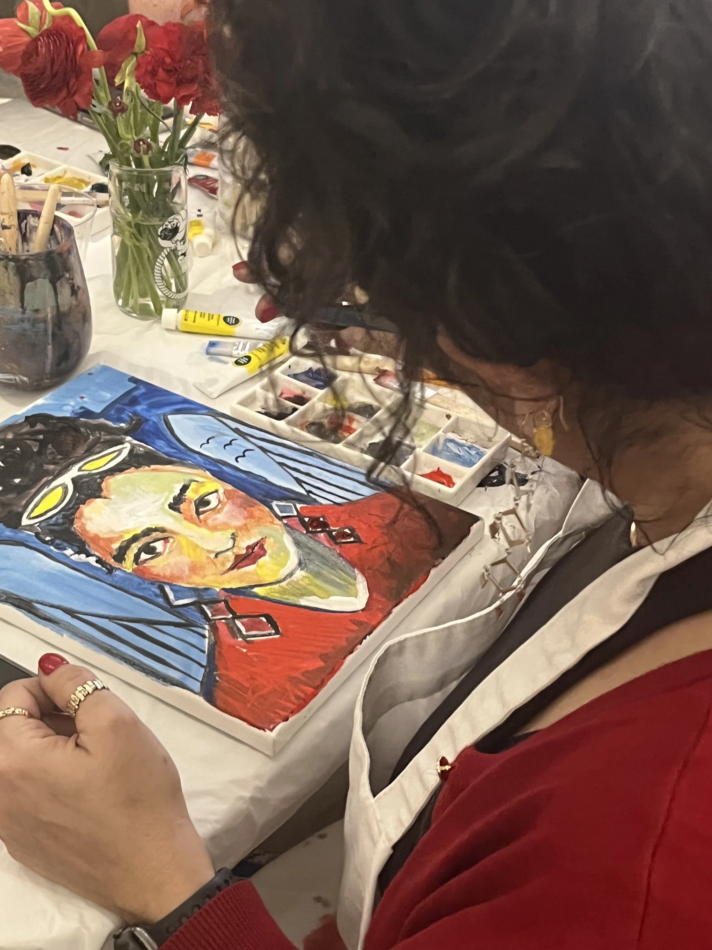 A woman with dark curly hair and red nail polish is painting a colorful portrait of a woman in a red dress on a canvas. She is seated at a table with art supplies, including tubes of paint, brushes, and a palette. There are red flowers in a glass vase in the background.