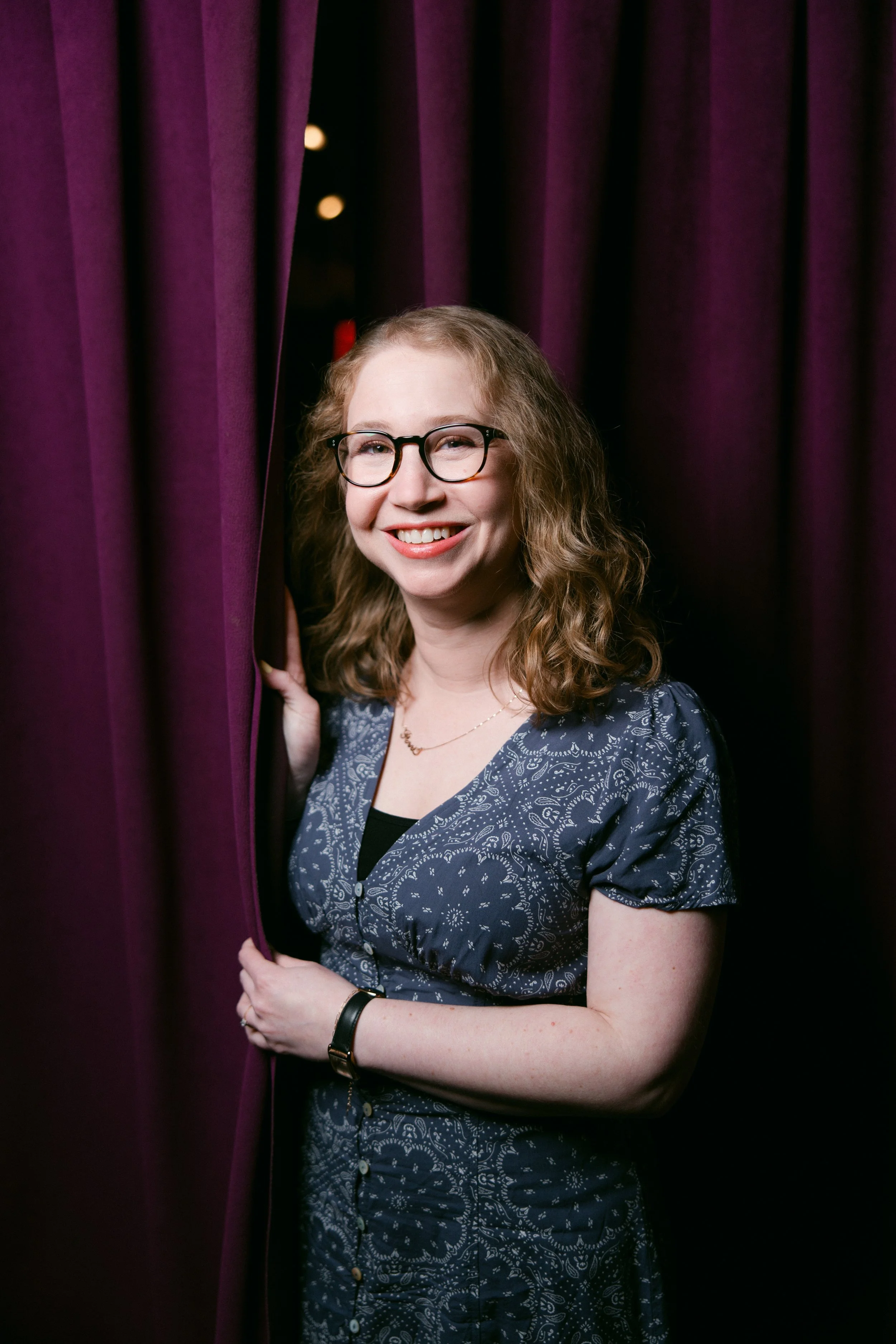 A woman with long, curly blonde hair, wearing glasses, a navy blue patterned dress, and a black watch, is smiling while peeking out from behind purple curtains.