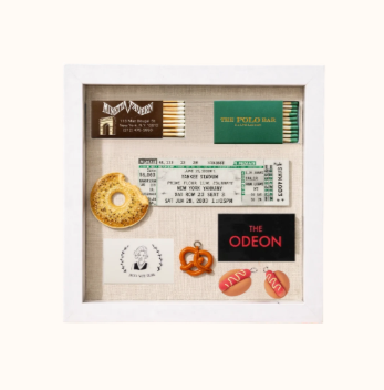 A shadow box with a movie ticket, a bag of The Polo Bar mints, a donut-shaped snack, a small pretzel, a black card for The Odeon, and two shrimp-shaped gummies.