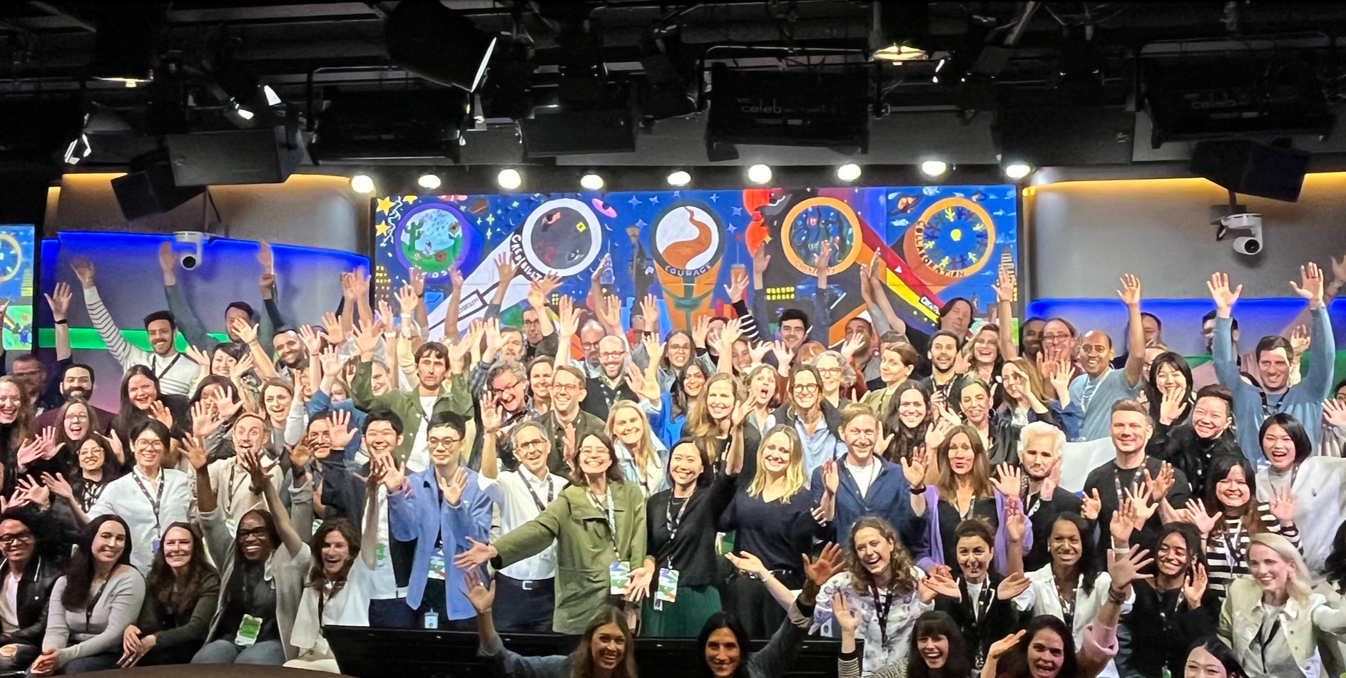 A large group of people gathered in front of a colorful digital backdrop, smiling and waving at the camera.