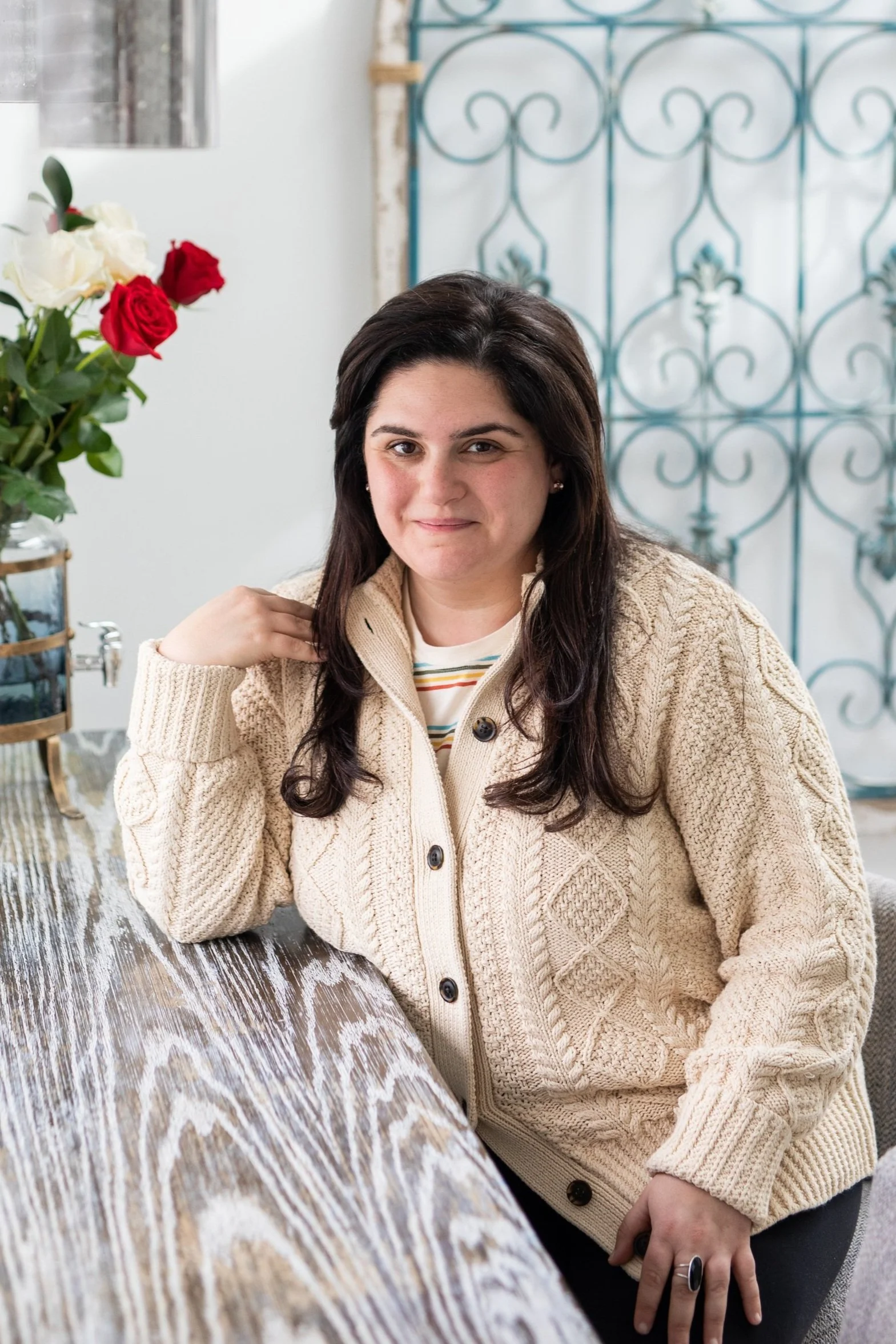 Olivia Howell sitting at a wooden table, wearing a cream-colored knit sweater, with a bouquet of red and white roses nearby.
