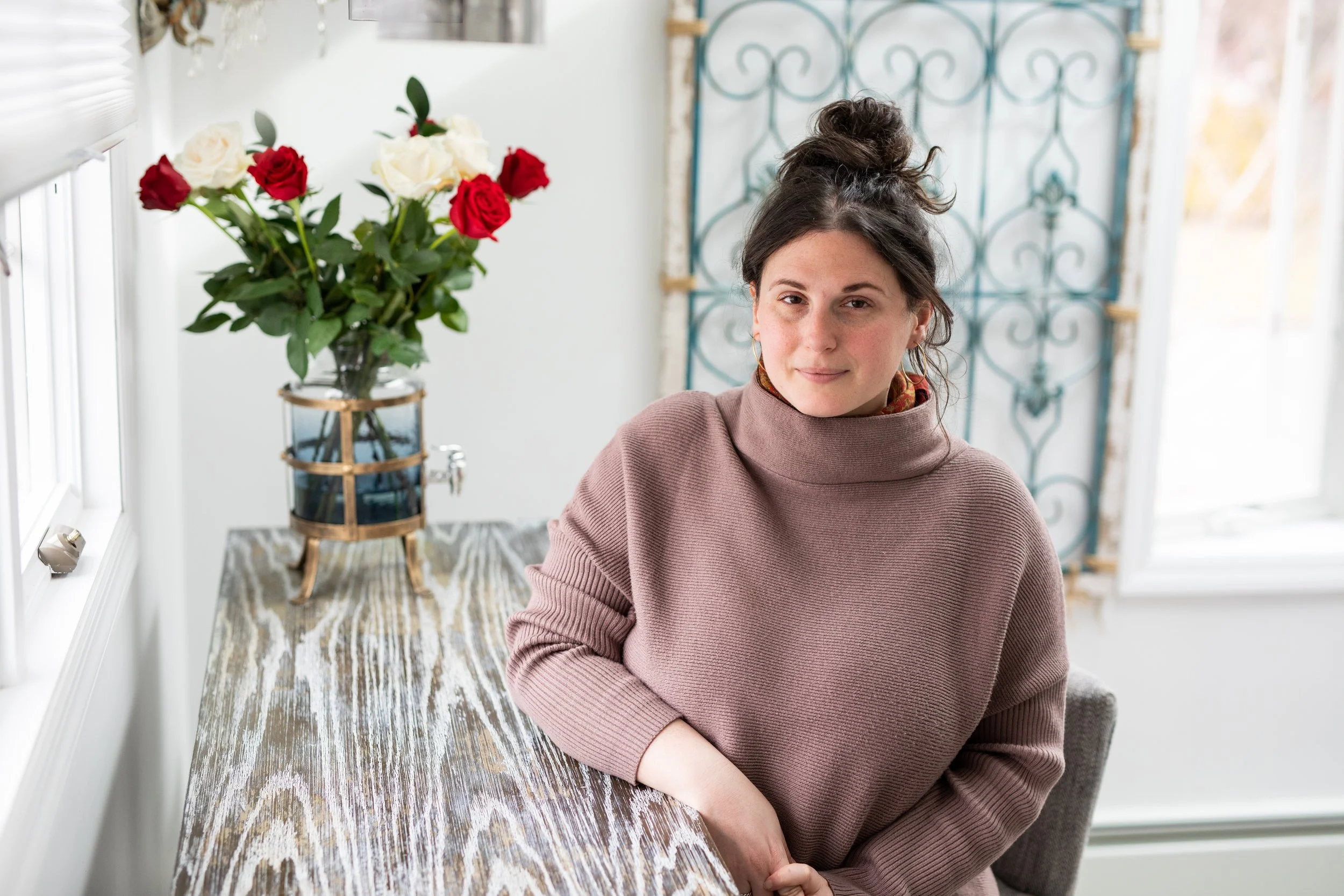 Genevieve Dreizen with dark hair in a bun, wearing a brown turtleneck, sitting at a wooden table with a glass dispenser and a bouquet of red and white roses.