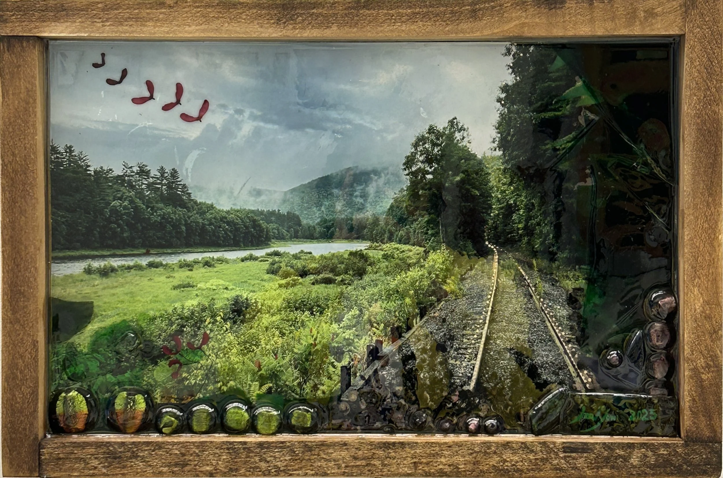 Framed landscape painting of a lush green countryside with a river, trees, hills, and train tracks, viewed through clear glass with some reflections.