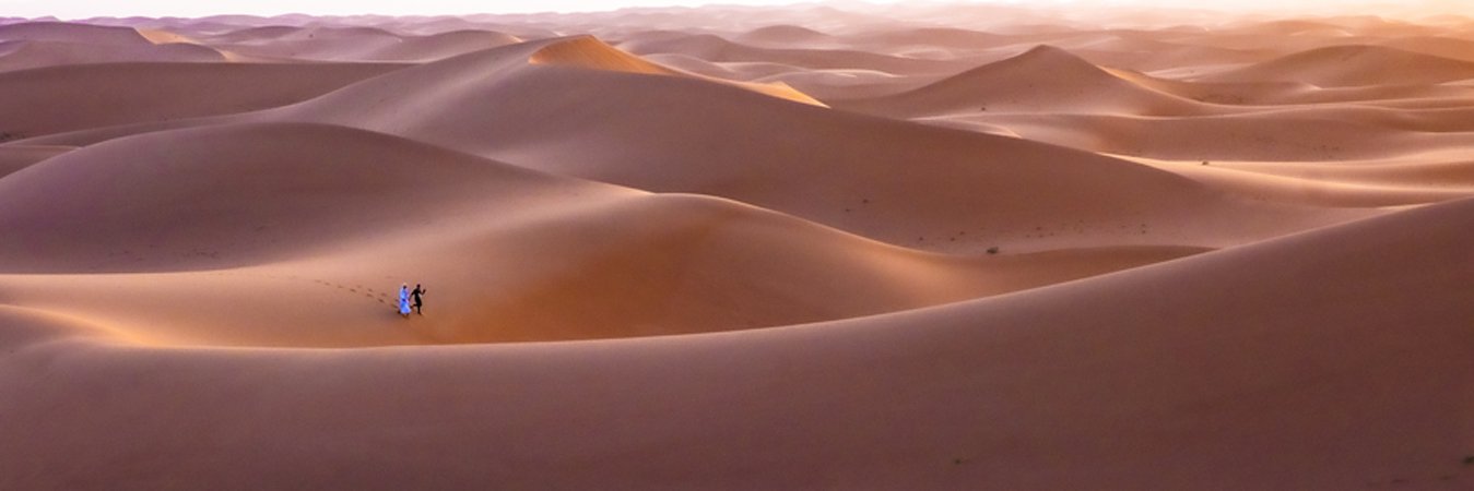 Two people walking on sand dunes in a desert at sunset.