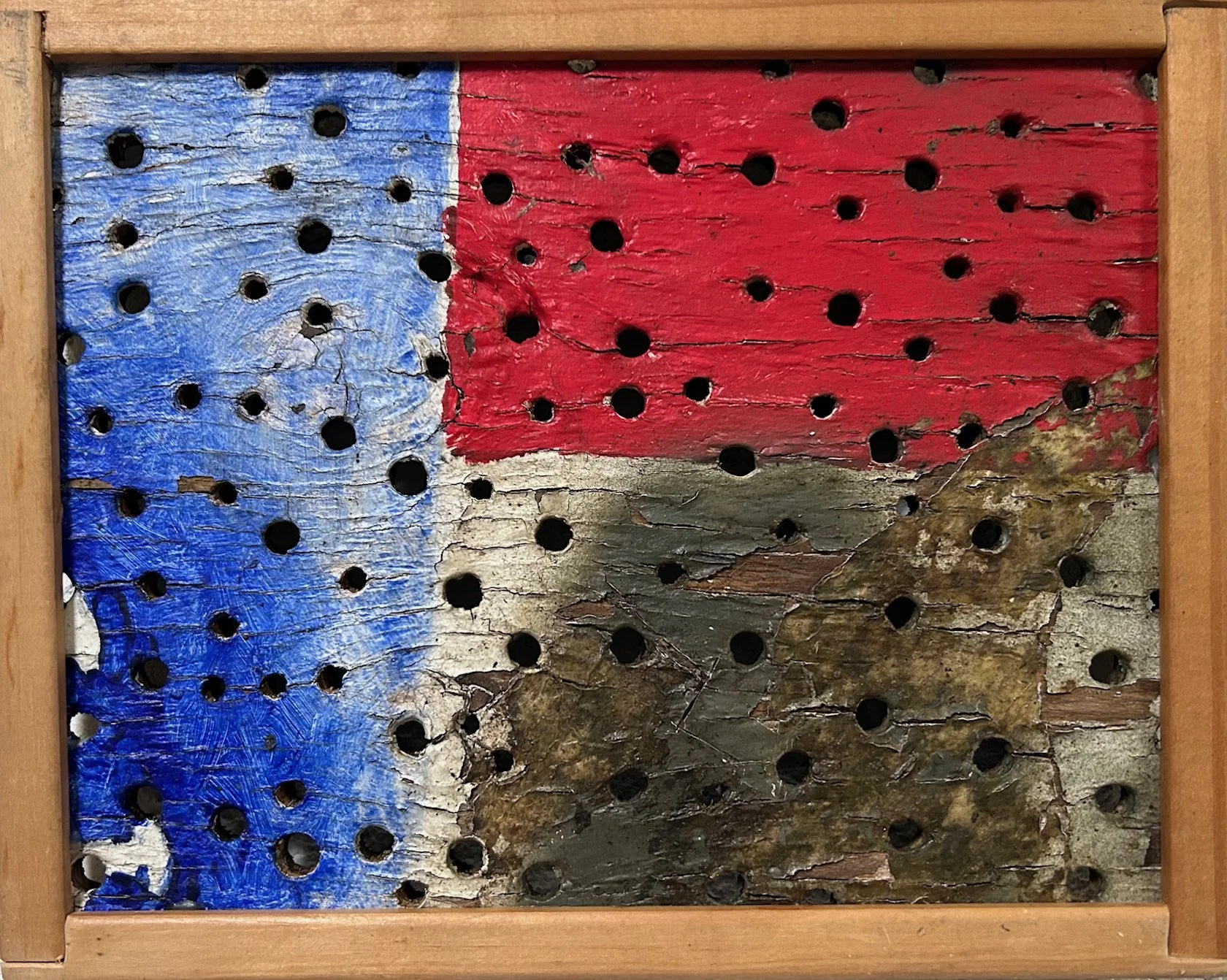A piece of old, weathered wood painted with the American flag, featuring a blue field with white stars in the bottom left, red stripes at the top, and distressed, peeling paint showing cracks and natural wood beneath.