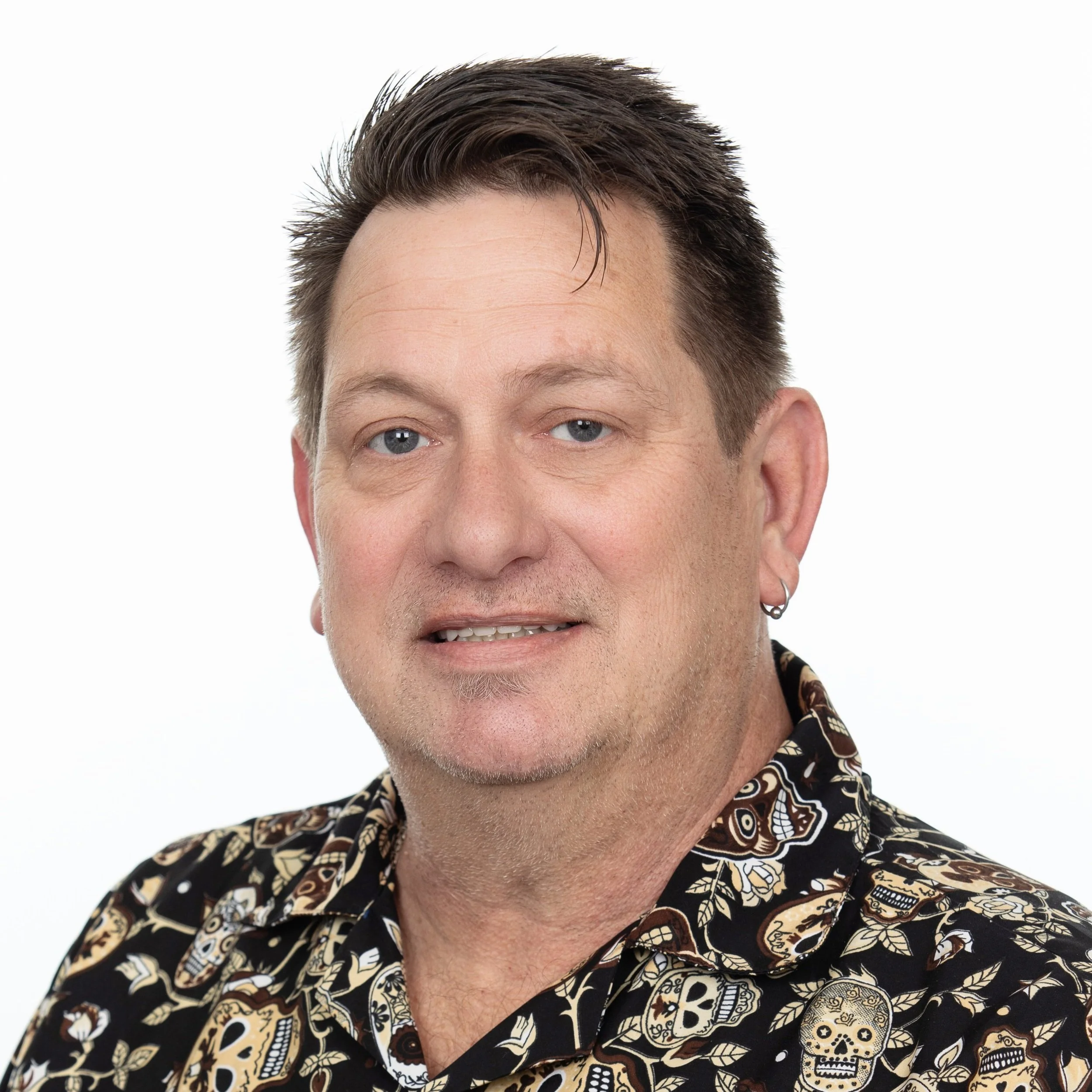 A man smiling with short brown hair wearing an earring with a black collared shirt with a pattern on it.