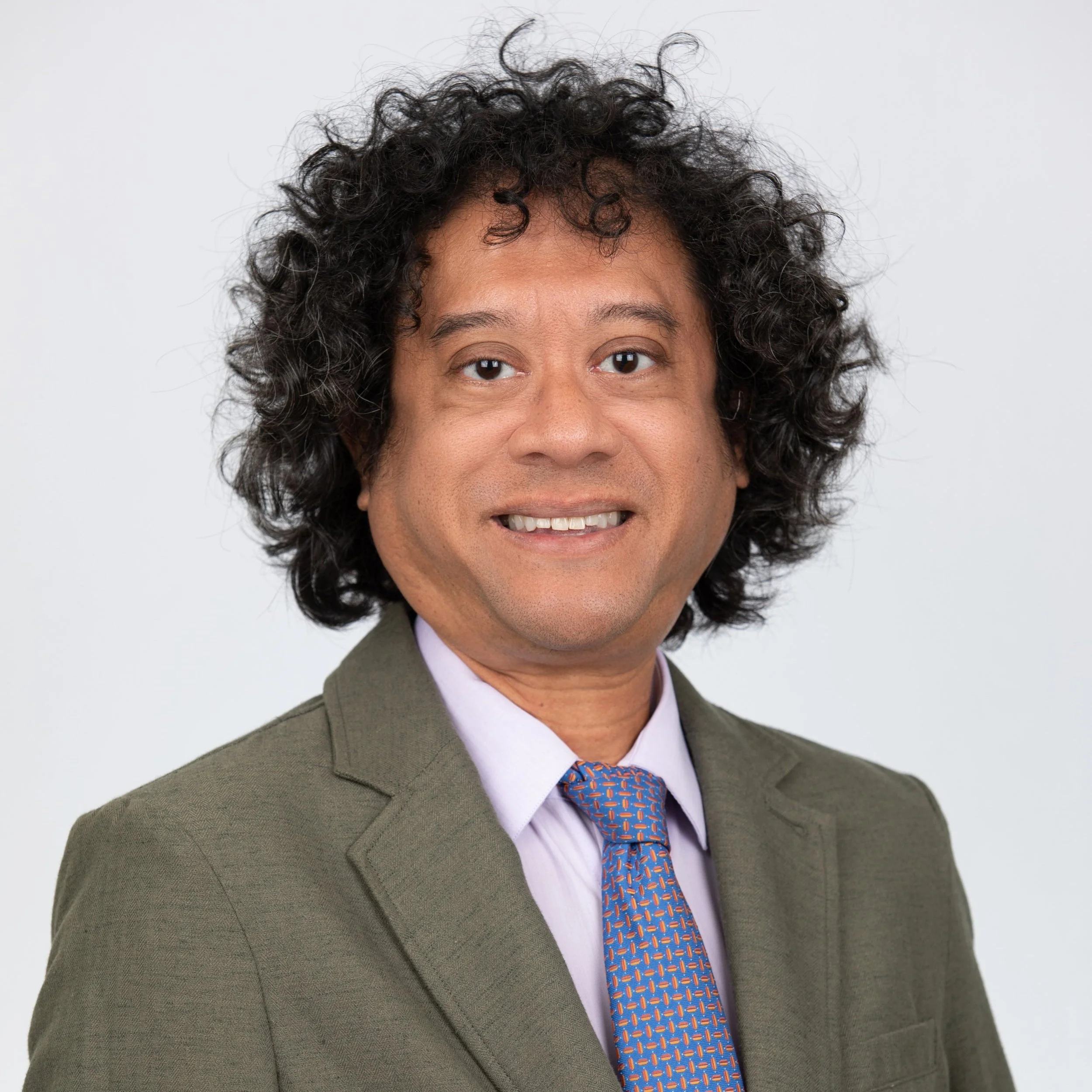 A man with tight curly dark hair wearing an olive green suit with a blue patterned tie.