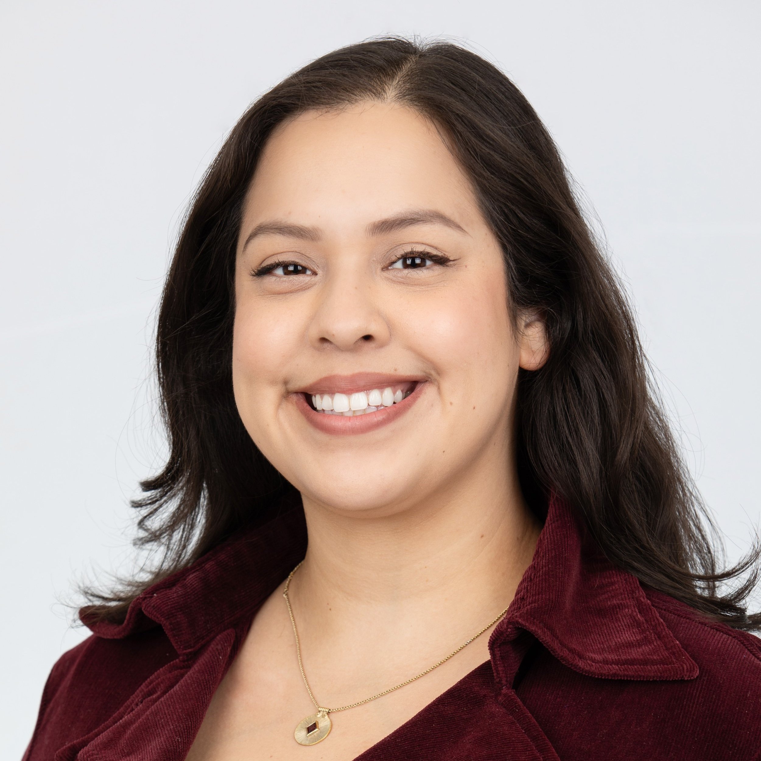 A woman smiling with shoulder length brown hair wearing a dark red button up shirt with a short gold necklace.