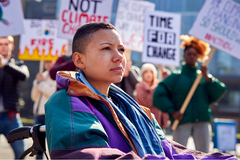 A woman in a wheelchair at an outdoor climate protest, wearing a colorful jacket and blue scarf, surrounded by people holding signs.