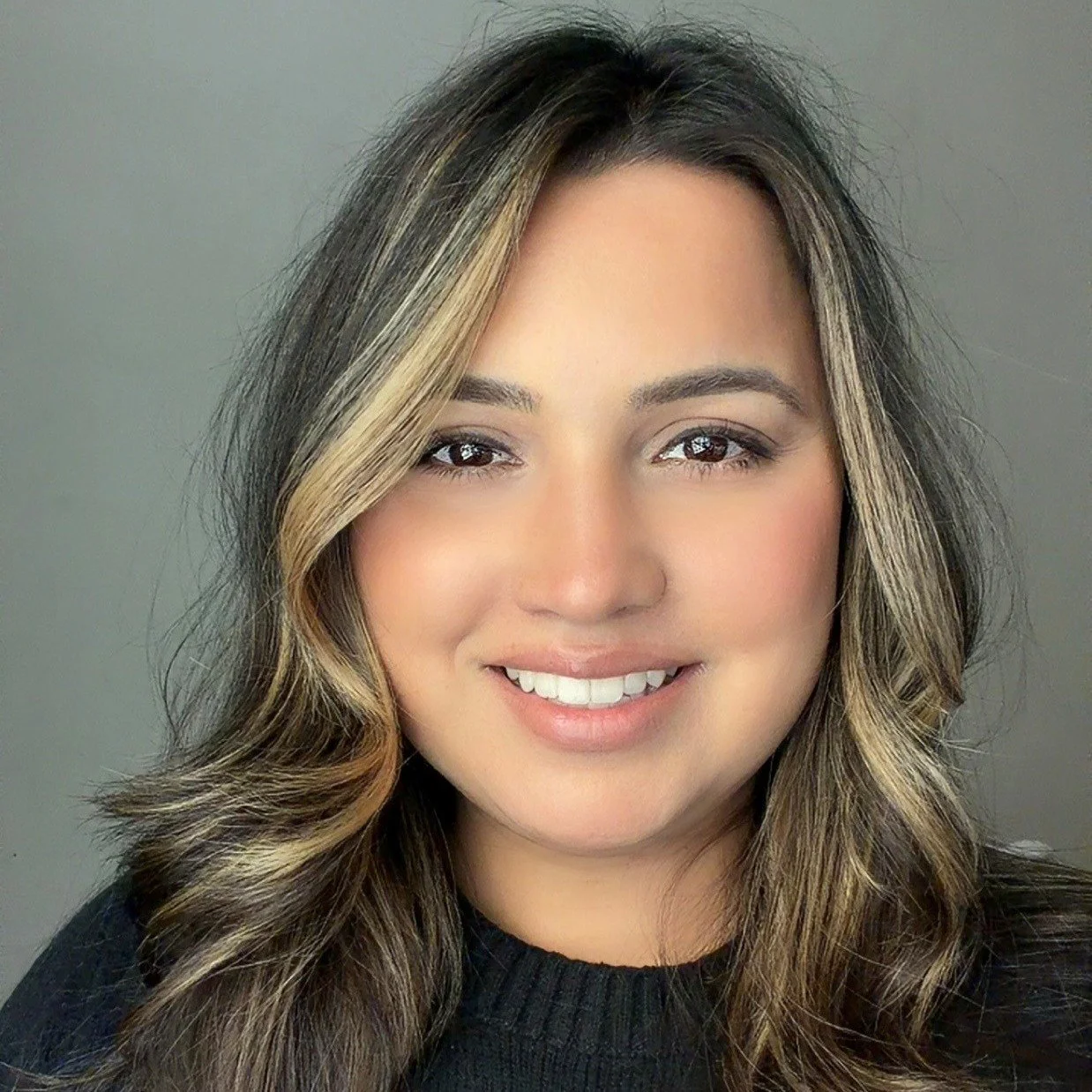 Smiling woman with long brown hair with blonde highlights, wearing black crew neck sweater.