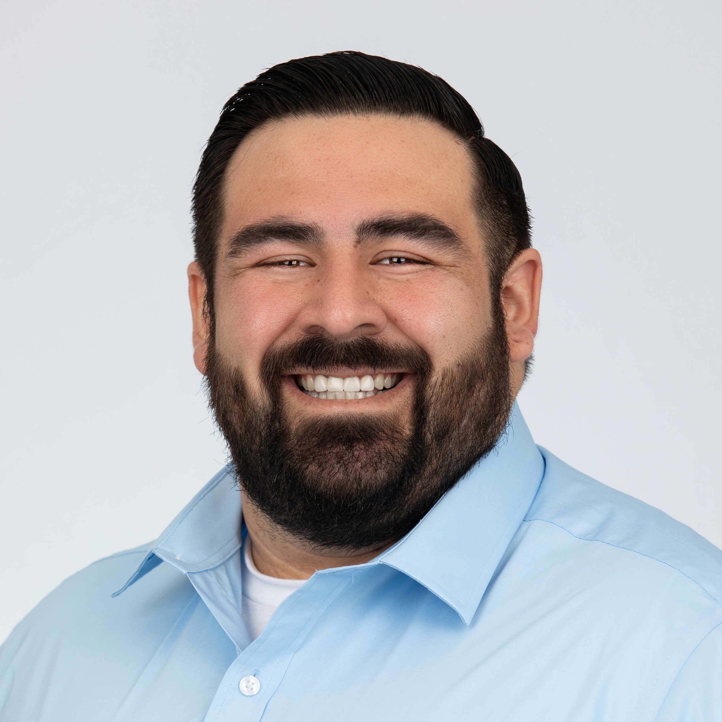 A man smiling with a dark brown beard and short combed dark hair with a light blue collared button up shirt with a white shirt underneath.